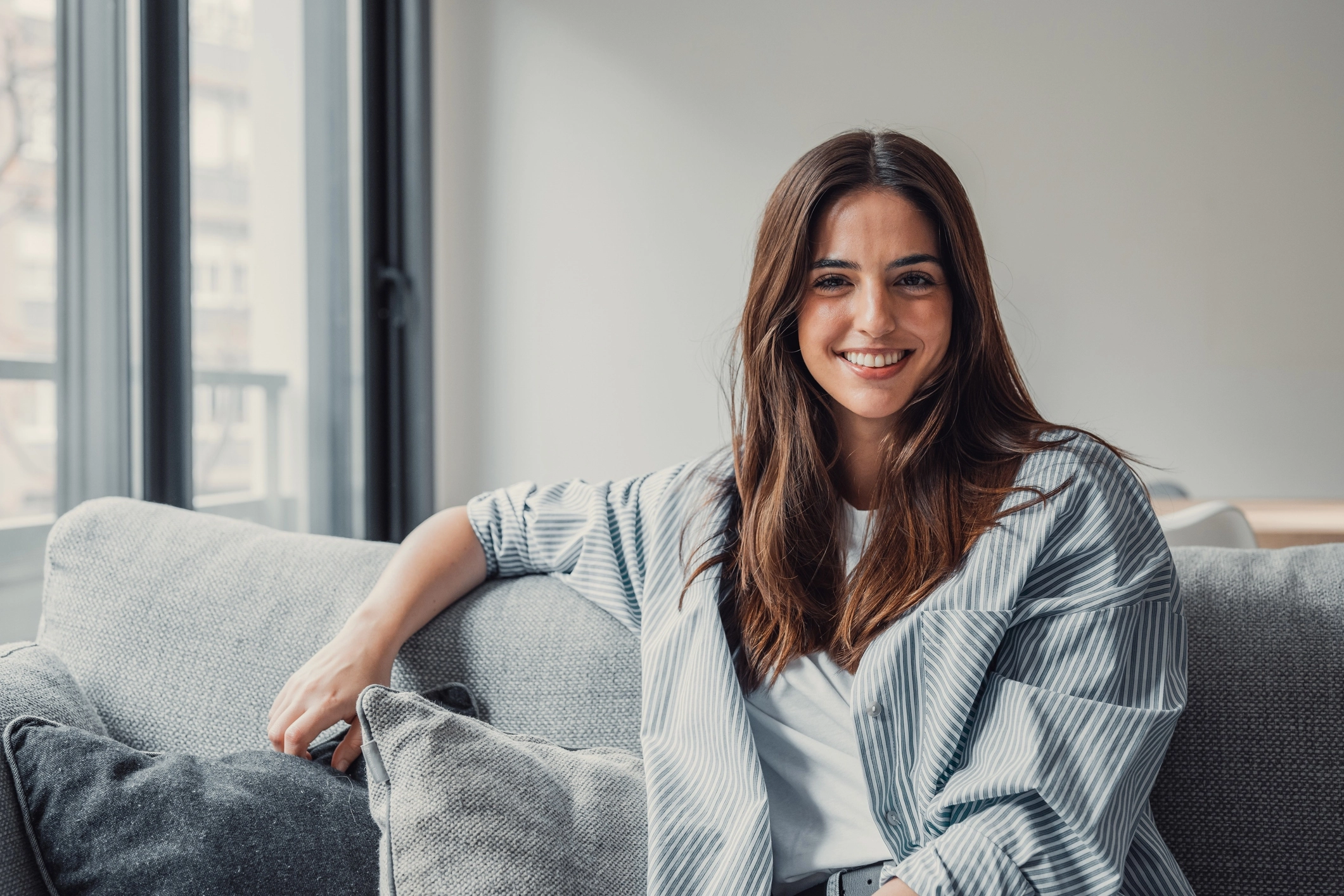 Happy attractive young adult woman girl sitting on sofa looking at camera in modern cozy home, smiling single 20s lady beautiful face relaxing on couch alone enjoy wellbeing posing for portrait